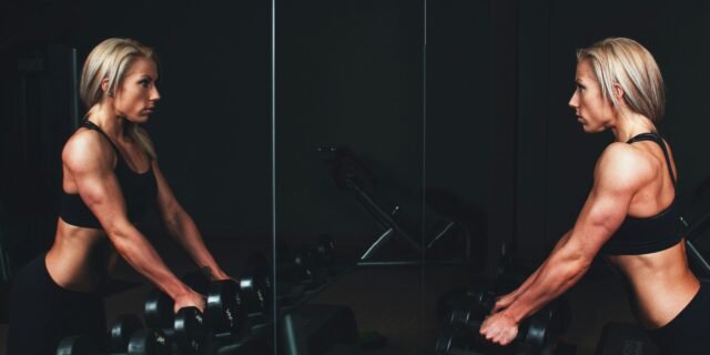 woman wearing black top top holding black dumbbells standing in front of mirror